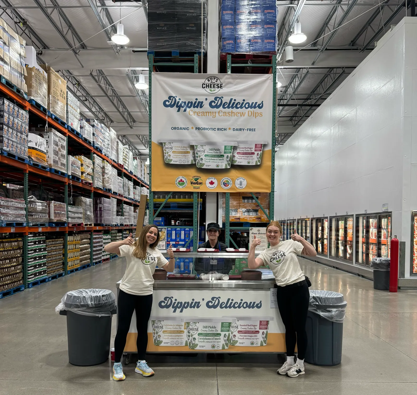 Nuts For Cheese roadshow station in a Costco warehouse: a Dippin Delicious hanging banner, a wrapped sampling cart and two team members in branded shirts